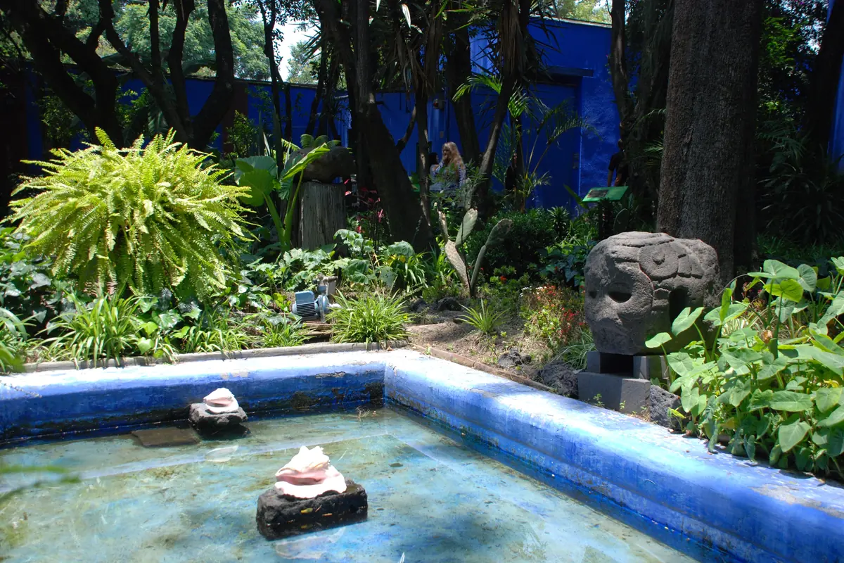 A shallow blue-tiled reflection pool in the garden of the Casa Azul, with shells in the water, ferns and a carved stone head at the edge.