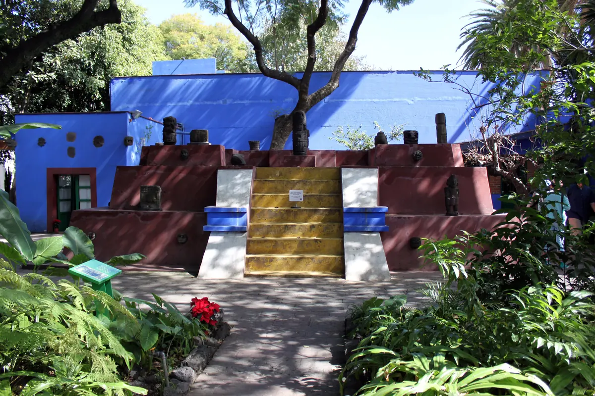 A stepped pyramid in red, yellow and white in the garden of the Casa Azul, with pre-Hispanic stone figures on its tiers and blue walls behind.
