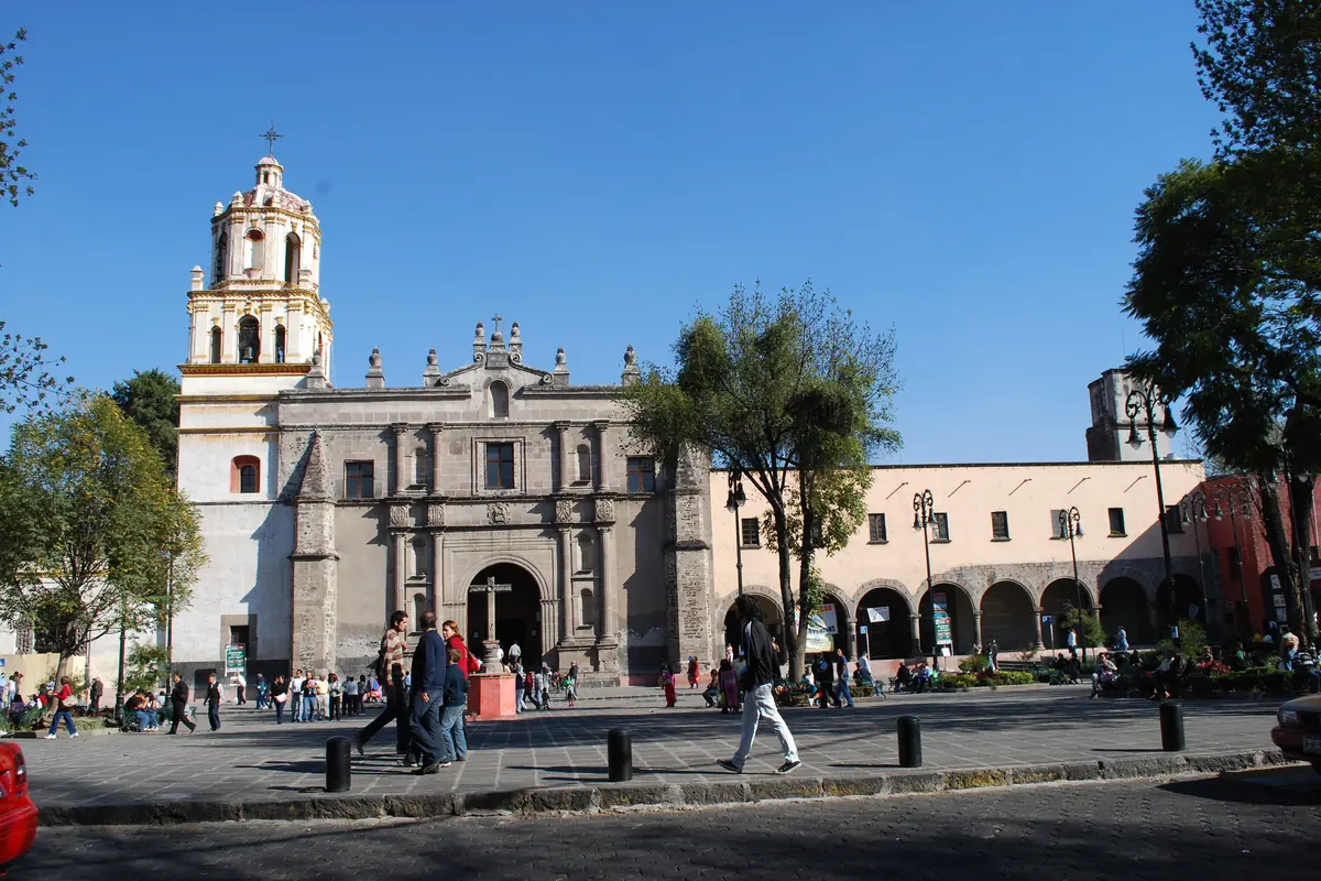 The stone facade and bell tower of the San Juan Bautista church on a sunny plaza in central Coyoacán.